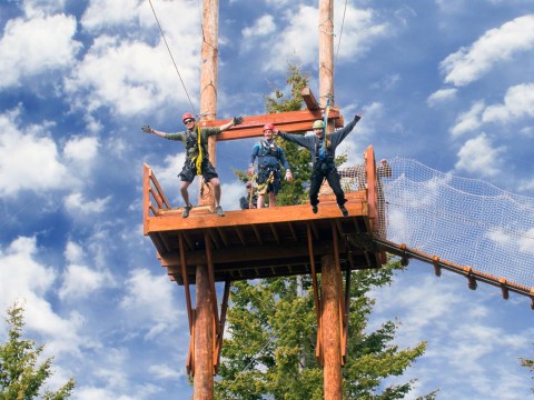 Three people in helmets jump from a high wooden platform against a cloudy sky.