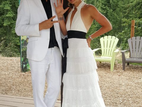 Couple in white formal attire posing outdoors with trees in the background.