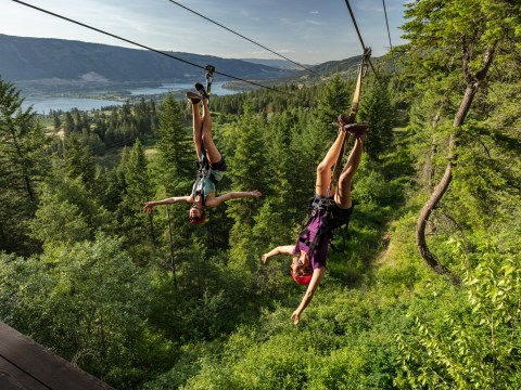 Two people ziplining upside down over a lush forest with a lake and mountains in the background.