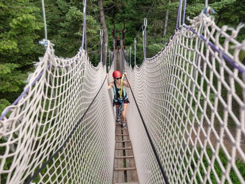 Person crossing a narrow rope bridge in a forest, wearing a helmet and safety harness.