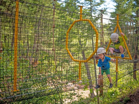 Two kids in helmets climb a rope course with yellow rings in a forest.