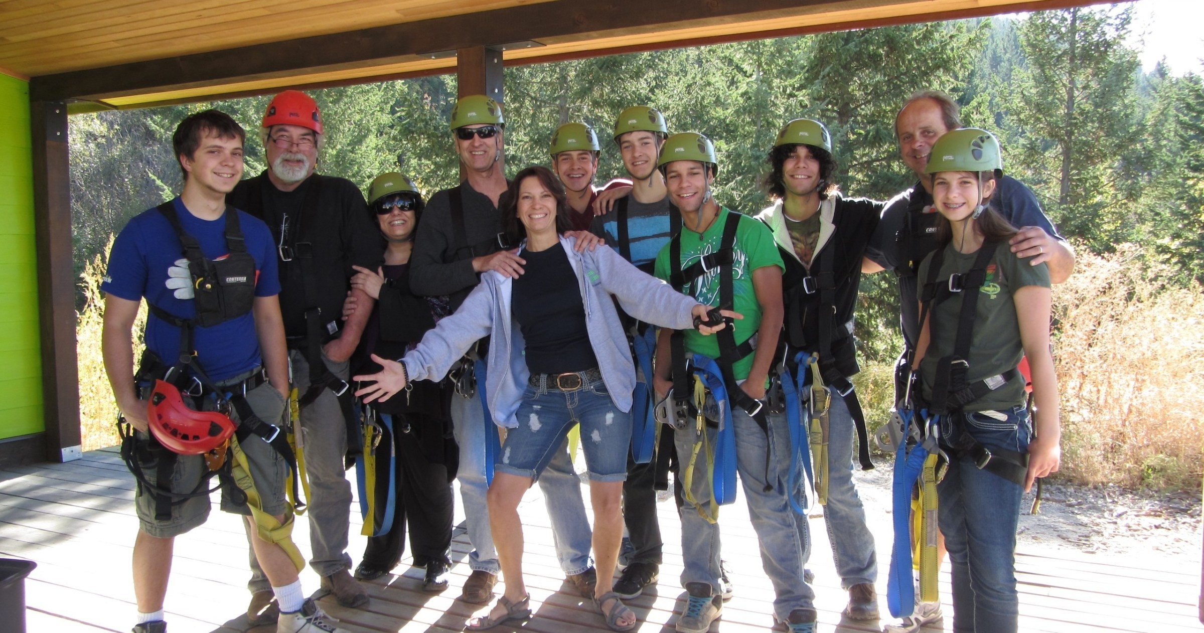 Group of people wearing helmets and harnesses, posing outdoors on a wooden deck with trees in the background.