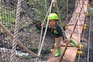 Child wearing a helmet and boots crossing a rope bridge in an adventure park.