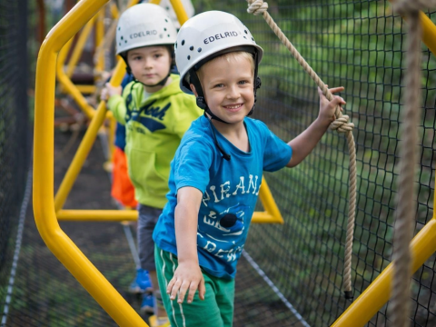 Two children wearing helmets navigate a rope bridge with netting beside them.