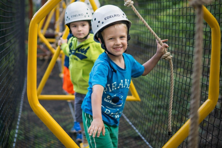 Two children wearing helmets navigate a rope bridge with netting beside them.